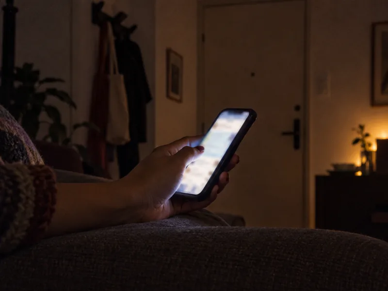 Mão segurando o celular aceso numa sala escura, no sofá, perto da porta de entrada